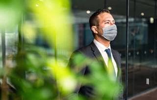 Man in a suit and wearing a mask standing next to a window