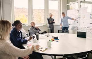 A group of people in an office watching a colleague present data on a whiteboard.