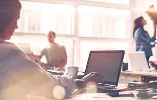 Photo over the shoulder of a woman working on a laptop in a busy office, with other professionals working on laptops in the bakground