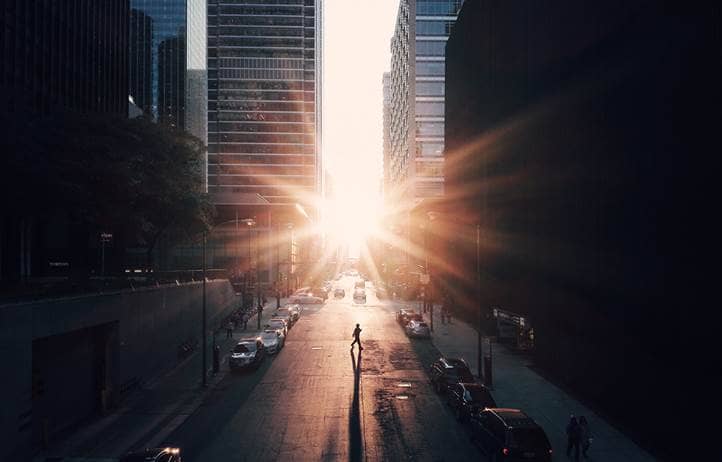 Aerial view of a city street bracketed by skyscrapers at sunset with the sun peaking between the buildings