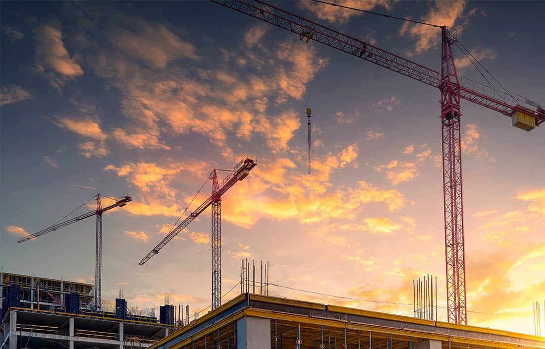 Construction of a commercial property with three high-rise cranes against the backdrop of a sunset.