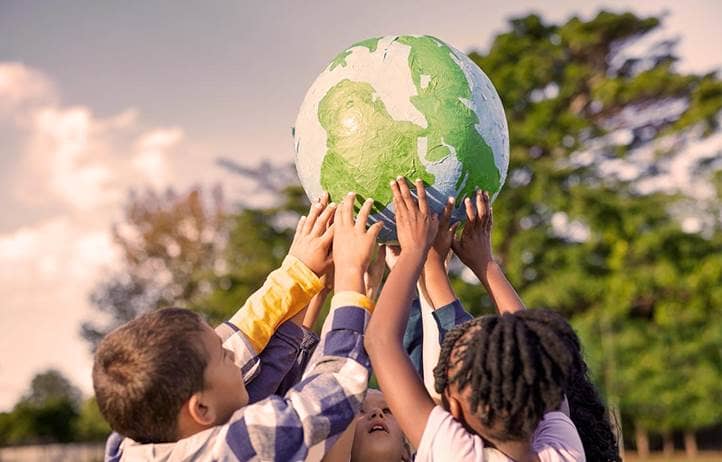 Group of children holding up a ball painted to look like the earth above their heads in a park with a tree and blue sky in the background