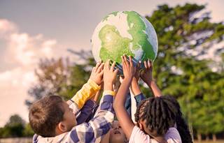 Group of children holding up a ball painted to look like the earth above their heads in a park with a tree and blue sky in the background