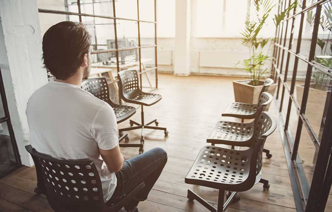 Man sitting in a chair in an office facing multiple empty chairs.